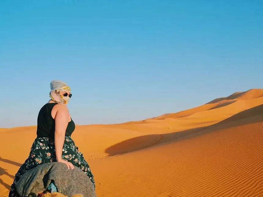 Voyageuse coiffée d’un foulard, à dos de dromadaire face aux dunes